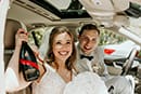 bride and groom sitting in getaway car holding bottle of champagne up