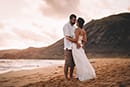 bride and groom standing on white sand beach with pink tones in the sky